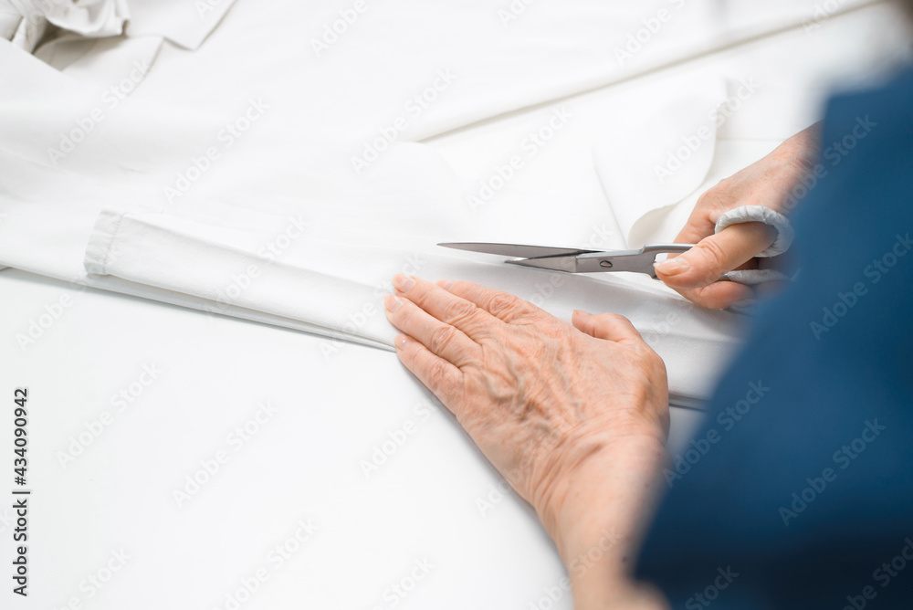 Elderly woman cutting white fabric with scissors, hands close-up. Senior woman dressmaker. Retired work or hobby at home. Elderly woman cutting white fabric with scissors, hands close-up. Senior woman dressmaker. Retired work or hobby at home.