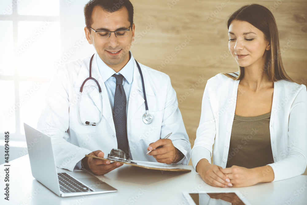 Doctor and patient discussing medical exam results while sitting at the desk in sunny clinic Doctor and patient discussing medical exam results while sitting at the desk in sunny clinic