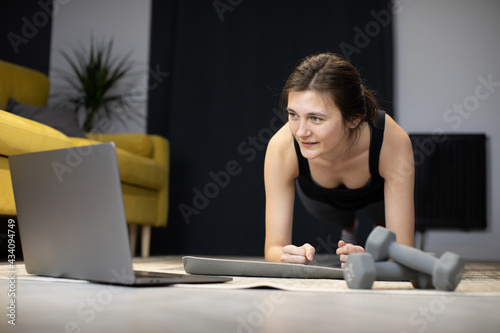 Smiling Girl stands in plank on floor of cozy living room looking at laptop counting seconds. Distance sports classes, working out. plank Challenge online concept. Young woman does abs exercise