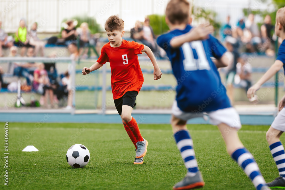 Young Boy Running Fast and Kicking Soccer Ball on Grass Pitch. Football Tournament For Youth School Teams. Group of Children in Jersey Unifrom Playing Sports. Football Fans and Parents in Background