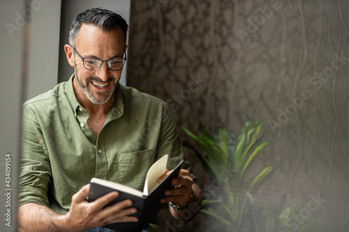 Mature man reading book by window, smiling wearing glasses, copy space