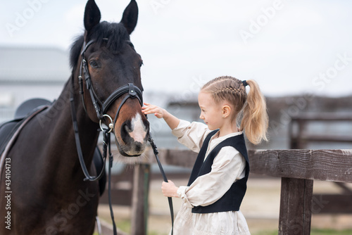 A 6-7 year old girl is stroking a horse and wants to feed him, holding a horse by the bridle. Equestrian sports for children
