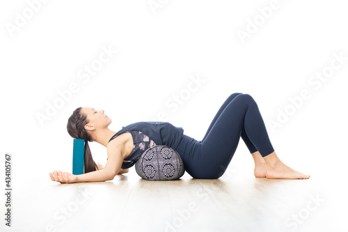 Restorative yoga with a bolster. Young sporty attractive woman in bright white yoga studio, lying on bolster cushion, stretching and relaxing during restorative yoga. Healthy active lifestyle