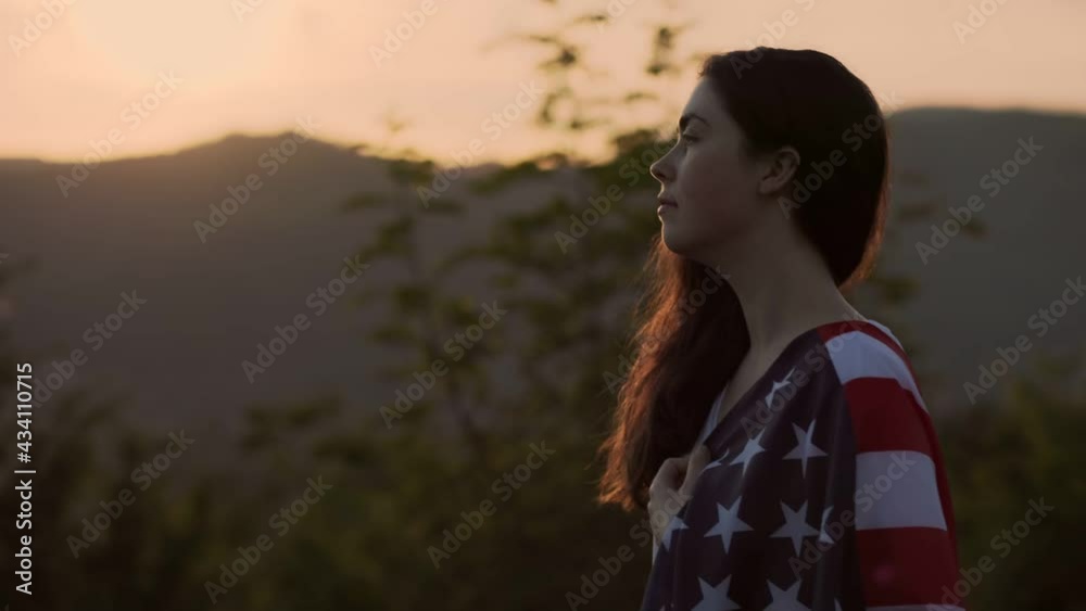 A young caucasian woman with an American flag draped over her shoulder, against the background of the sunset sky. Slow motion. Side view. Dolly shot. The concept of patriotism A young caucasian woman with an American flag draped over her shoulder, against the background of the sunset sky. Slow motion. Side view. Dolly shot. The concept of patriotism