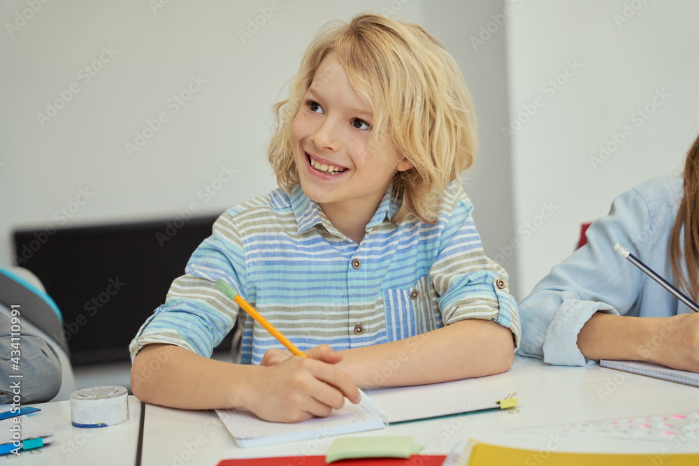 Happy little schoolboy smiling away, holding pencil and listening while studying, sitting at the table in elementary school classroom Happy little schoolboy smiling away, holding pencil and listening while studying, sitting at the table in elementary school classroom