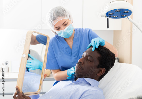 Professional cosmetologist examining male client face before procedure in esthetic clinic. Focused man looking at mirror