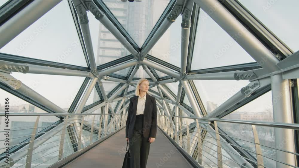 Front-view drone slowmo of confident middle-aged businesswoman in black suit walking along long empty glass bridge of classy office building Front-view drone slowmo of confident middle-aged businesswoman in black suit walking along long empty glass bridge of classy office building