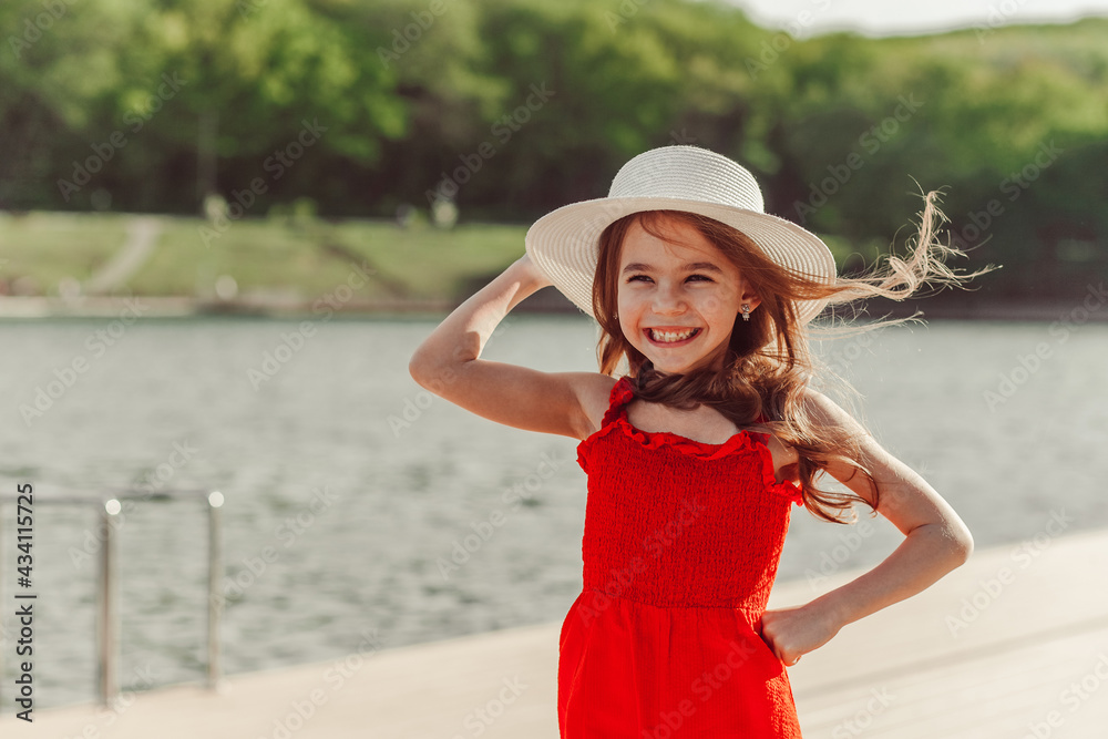 Happy girl in a red jumpsuit and white hat on the waterfront. Enjoyment of nature, freedom, life after quarantine.