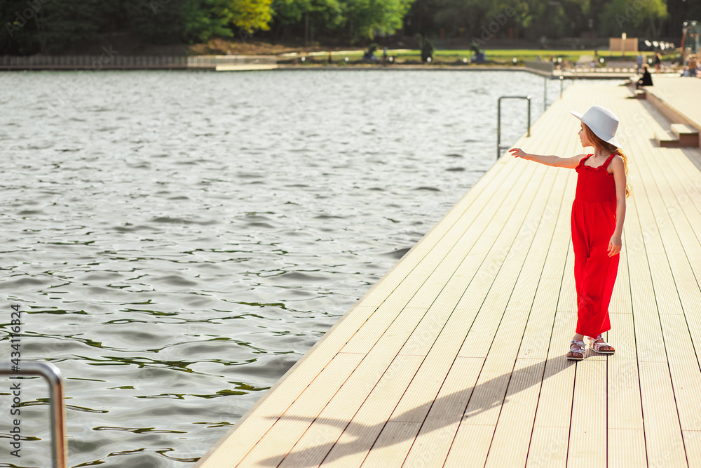 Fototapeta premium Happy girl in a red jumpsuit and white hat on the waterfront. Enjoyment of nature, freedom, life after quarantine.