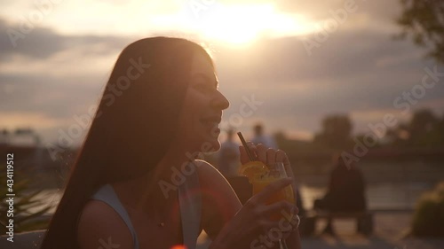 Smiling young woman drinks orange juice  at sunset outdoor photo