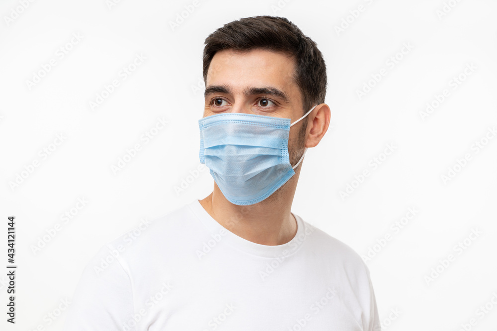 Young handsome man wearing white t-shirt and medical mask, standing isolated on gray background, looking away
