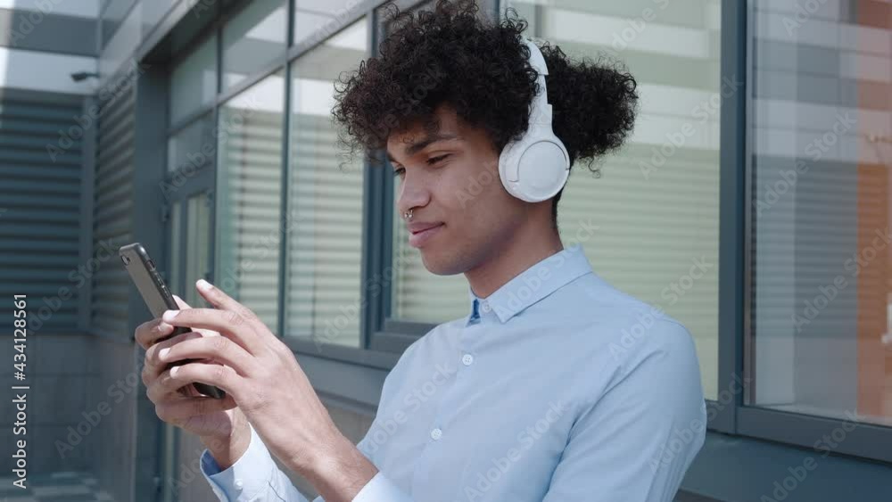 Ethnic man in headphones looking at smartphone near building in town Ethnic man in headphones looking at smartphone near building in town