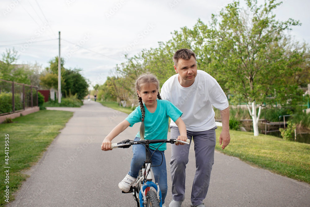 Father teaching her little daughter to ride a bicycle, happy summer in village, healthy lifestyle, childhood, fatherhood and upbringing, concept for father day Father teaching her little daughter to ride a bicycle, happy summer in village, healthy lifestyle, childhood, fatherhood and upbringing, concept for father day