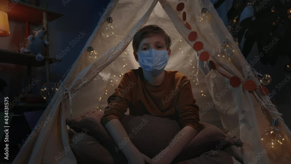 Portrait of young boy in protective medical mask sitting in decorative makeshift hut at home in evening. Teen looking to camera.Concept of quarantine, illness. Portrait of young boy in protective medical mask sitting in decorative makeshift hut at home in evening. Teen looking to camera.Concept of quarantine, illness.