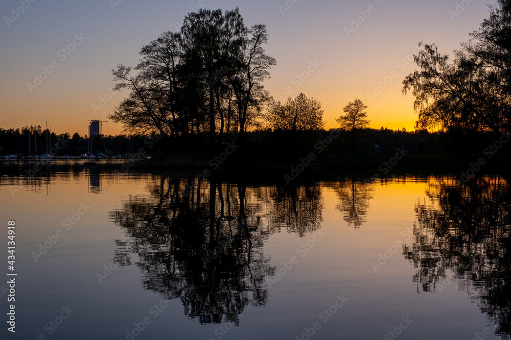 A beautiful silhouette of a large tree on an island casting reflection on the water during sunset.