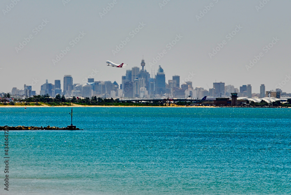 Fototapeta premium Southern view of the Sydney skyline as a plane takes off on a hazy summer day, seen from the shoreline of Botany Bay in New South Wales, Australia.