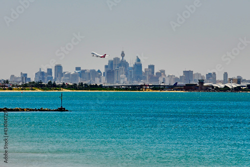 Southern view of the Sydney skyline as a plane takes off on a hazy summer day, seen from the shoreline of Botany Bay in New South Wales, Australia.