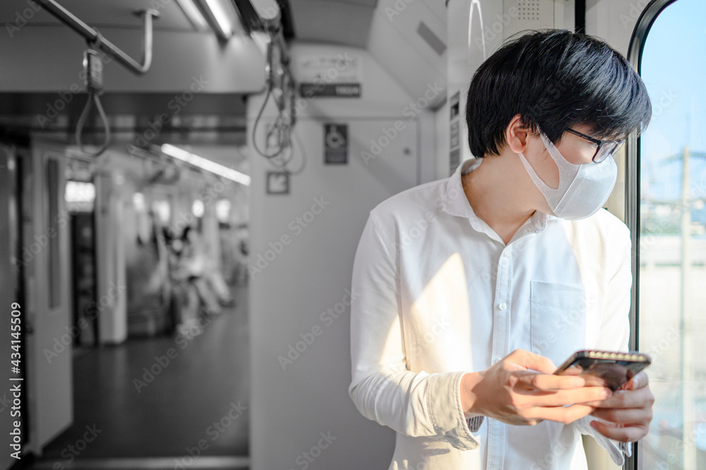 Asian man wearing face mask using smartphone on skytrain or urban train. Coronavirus (COVID-19) infection prevention in public transportation. Social distancing for pandemic protection