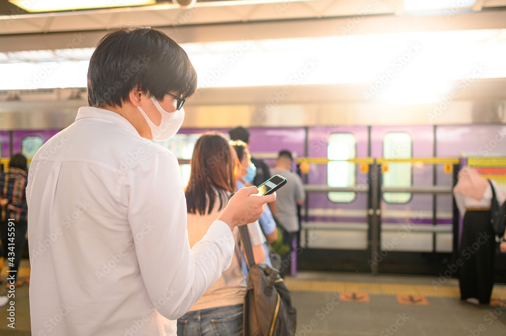 Asian man wearing face mask using smartphone at skytrain station platform. Coronavirus (COVID-19) infection prevention in public transportation. Health awareness for pandemic protection Asian man wearing face mask using smartphone at skytrain station platform. Coronavirus (COVID-19) infection prevention in public transportation. Health awareness for pandemic protection