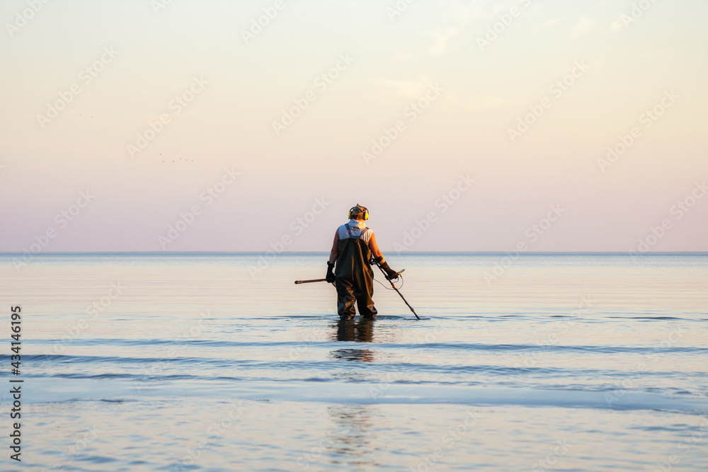 A man with a metal detector and scoop searches for treasures in the sea.