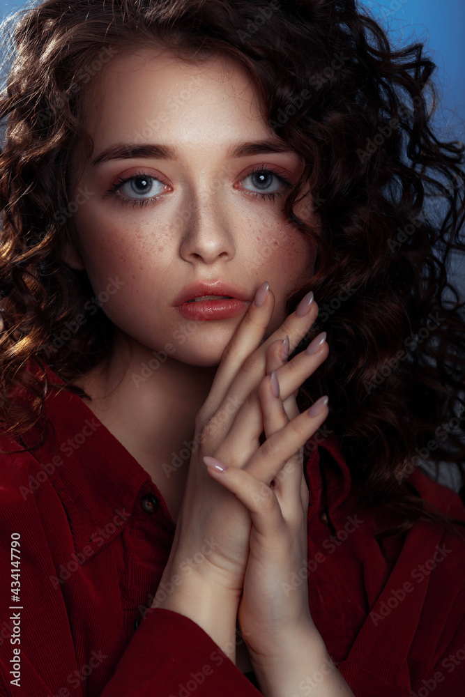 Attractive brunette with long curly hair. Female fashion model with wavy hairstyle in a red shirt posing on a blue background Attractive brunette with long curly hair. Female fashion model with wavy hairstyle in a red shirt posing on a blue background