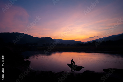 Evening landscape mountains river blue sky There was a thin orange cloud after sunset. And there is a man's shadow sitting on the rocks by the river Give you a feeling of loneliness