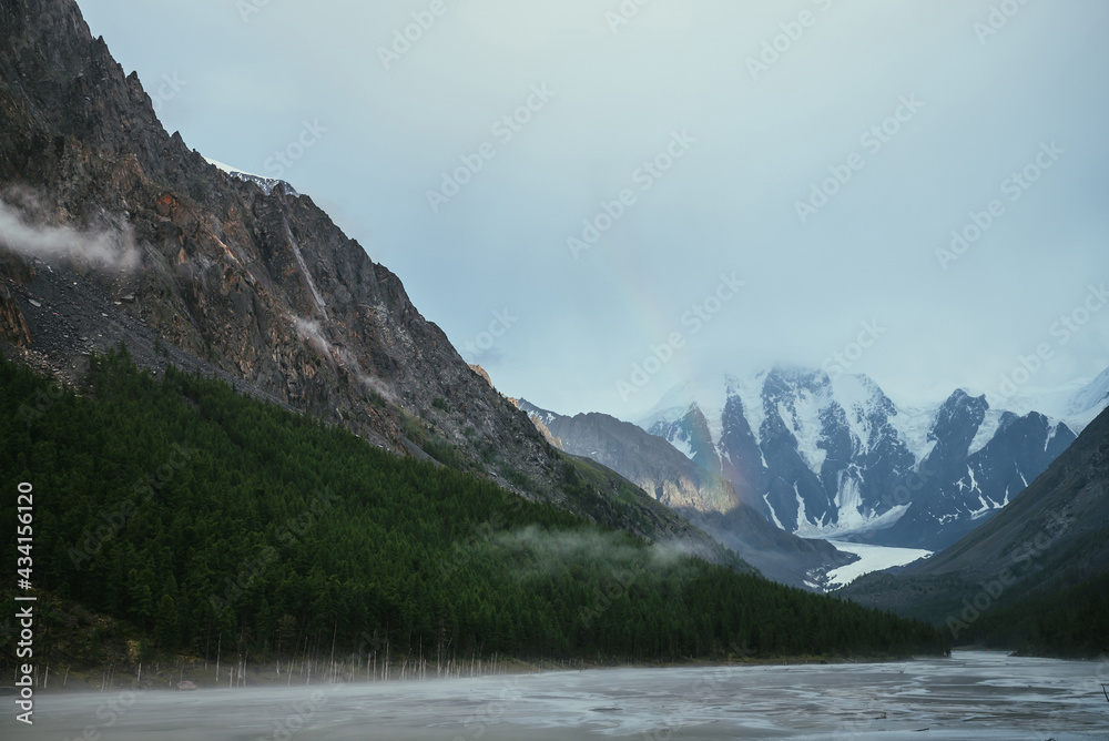 Scenic alpine landscape with mountain streams in fog and snowy mountains with rainbow under cloudy sky. Atmospheric scenery with shallow mountain lake, sunlight on rocks and low clouds in valley.