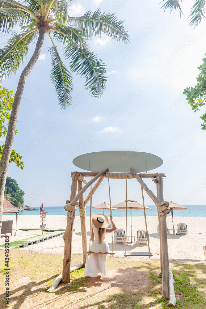 Back View of Travel Woman in White Dress and Straw Hat Sitting on Wooden Swing, Tropical Palms and Sandy Beach with Blue Sea on Background. Female Tourist Leisure in Resort on Phuket Island, Thailand