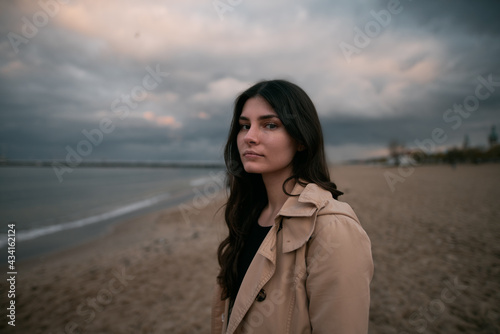 Portrait of long brown haired girl standing alone on the sand beach during cloudy weather. Spring or autumn vacation on the seaside concept. Close up portrait of caucasian woman.