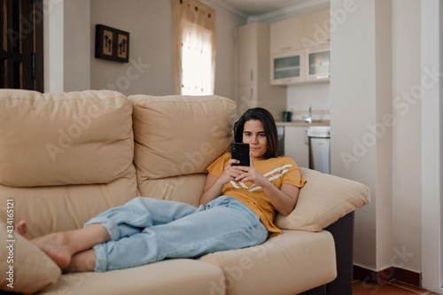 Young female text messaging on cellphone while lying down on couch in living room