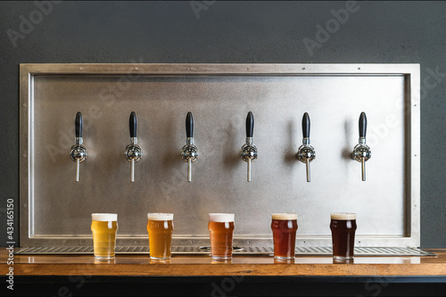 Different types of beer with foam in glass jugs against row of taps in bar on gray background photo