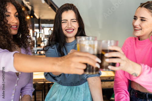 Joyful young multiethnic female best friends in stylish colorful outfits smiling and clinking glasses of refreshing juice and coke in modern bar