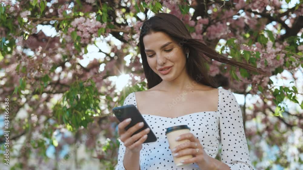 Woman with charming Smile walking in the Park and Using Mobile Phone Wearing Stylish Outfit. Girl Using Smartphone and Drinking Coffee. Communication, online shopping, social network concept Woman with charming Smile walking in the Park and Using Mobile Phone Wearing Stylish Outfit. Girl Using Smartphone and Drinking Coffee. Communication, online shopping, social network concept