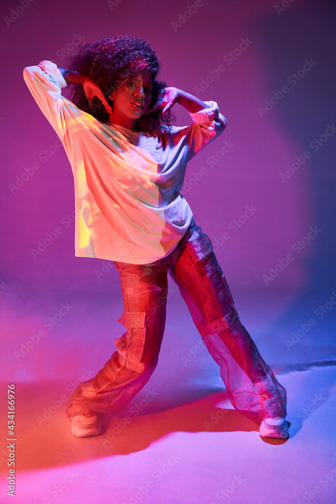 Full length fit young African American female dancer in loose informal wear touching curly hair and looking at camera while dancing in dark studio in neon lights Full length fit young African American female dancer in loose informal wear touching curly hair and looking at camera while dancing in dark studio in neon lights
