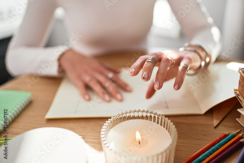 Crop unrecognizable astrologist diving future at desk with paper album and flaming candle in house