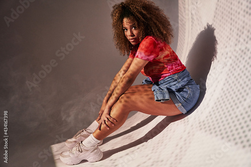 Side view full body gorgeous confident African American female in shorts sitting on curvy wall and looking at camera photo