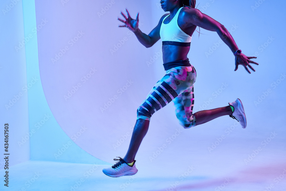 Side view of determined African American female athlete jumping while running and looking forward during training on blue background Side view of determined African American female athlete jumping while running and looking forward during training on blue background