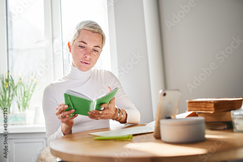 Adult female astrologist reading notepad at desk with smartphone and books at home in sunlight