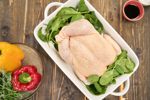 Top view of whole uncooked poultry with fresh spinach foliage in baking dish between peppers and soy sauce on table