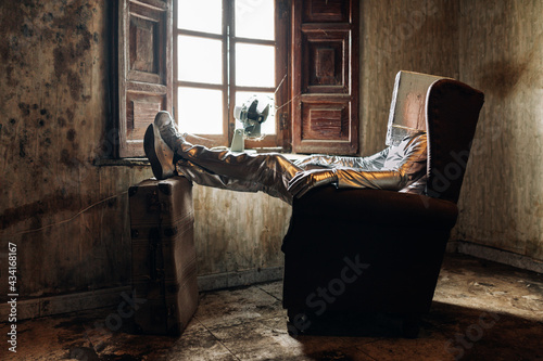 Side view unrecognizable person wearing protective silver suit with box on head resting on armchair in shabby room in abandoned house photo