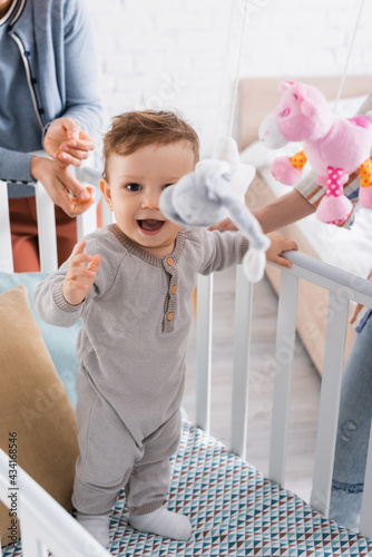 excited infant boy in baby crib with hanging soft toys on blurred foreground
