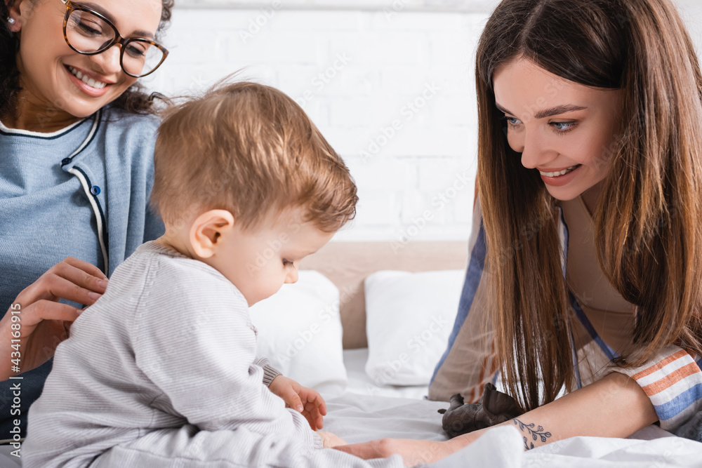interracial women looking at infant boy sitting on bed interracial women looking at infant boy sitting on bed