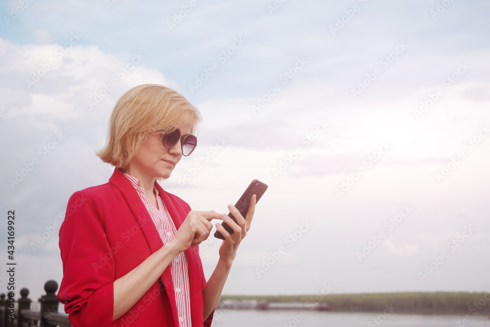 Исходный текст.Happy blonde caucasian girl in sunglasses and a pink jacket on a background of river shore. She holds a phone. Business woman concept..