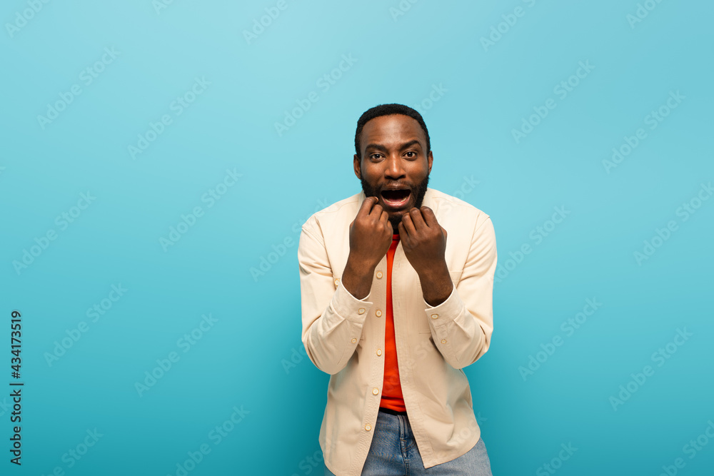 shocked african american man holding hands near face while looking at camera on blue background shocked african american man holding hands near face while looking at camera on blue background