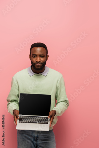 african american man looking at camera while holding laptop with blank screen on pink background