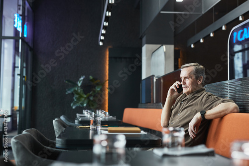 Confident senior Caucasian manager in brown shirt sitting in relaxed pose on bistro cafe seat and talking on phone