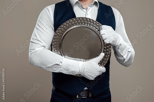 A male waiter in a white shirt and white gloves, standing with a silver tray, hugs it to his chest. The concept of serving staff, serving customers in a restaurant.