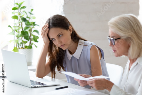 Unhappy young female employee bored with middle-aged colleague discuss paper document at meeting. Tired woman worker bothered annoyed by senior coworker lecture about paperwork at team briefing.