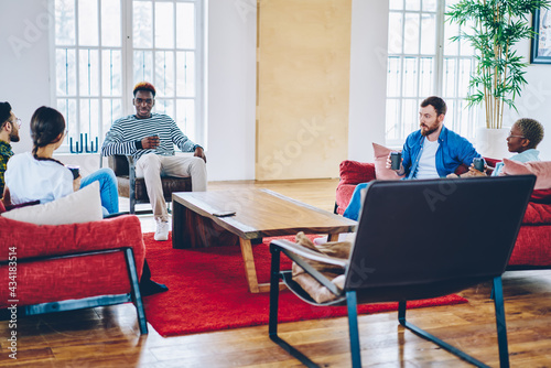 Group of multiracial people talking with friends sitting on comfortable red sofa enjoying coffee break in stylish home interior, young trendy people communicating in loft hostel apartment