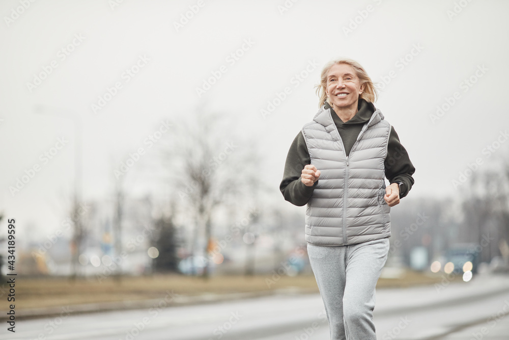 Positive energetic senior Caucasian sportswoman in warm vest and gray pants jogging along road while training in winter Positive energetic senior Caucasian sportswoman in warm vest and gray pants jogging along road while training in winter